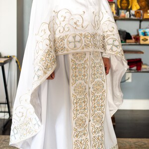 May include: White liturgical vestment with gold and blue embroidery. The vestment features intricate floral patterns and crosses. The fabric is smooth and silky.