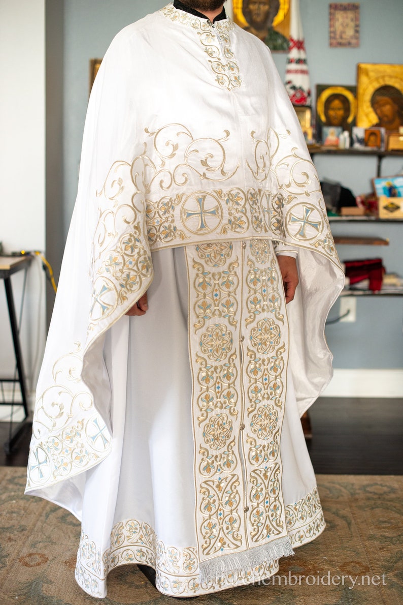 May include: White liturgical vestment with gold and blue embroidery. The vestment features intricate floral patterns and crosses. The fabric is smooth and silky.