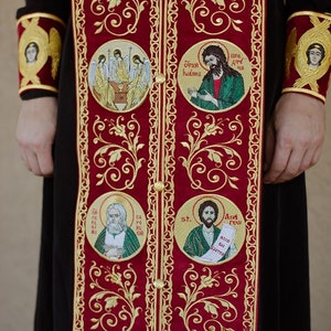 Pode incluir: Uma veste bordada em vermelho e dourado com m&uacute;ltiplos &iacute;cones religiosos, incluindo uma representa&ccedil;&atilde;o de Jesus Cristo, S&atilde;o Andr&eacute; e S&atilde;o Tikhon. A veste &eacute; decorada com padr&otilde;es florais intrincados.
