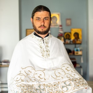 May include: A man wearing a white robe with gold and blue embroidery. The robe has a high collar and is decorated with intricate patterns. The man is looking at the camera with a serious expression.