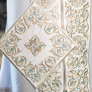 May include: White fabric with intricate gold and blue embroidery. The design features a repeating pattern of stylized flowers and leaves.