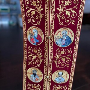 Pode incluir: Um tecido de veludo vermelho com bordados dourados. O tecido apresenta um padr&atilde;o repetitivo de desenhos florais e imagens circulares de figuras religiosas. O texto "&Epsilon;ὐ&lambda;&omicron;&gamma;&eta;&tau;ὸ&sigmaf; ὁ &Theta;&epsilon;ὸ&sigmaf; ἡ&mu;ῶ&nu;" est&aacute; bordado em ouro no tecido.