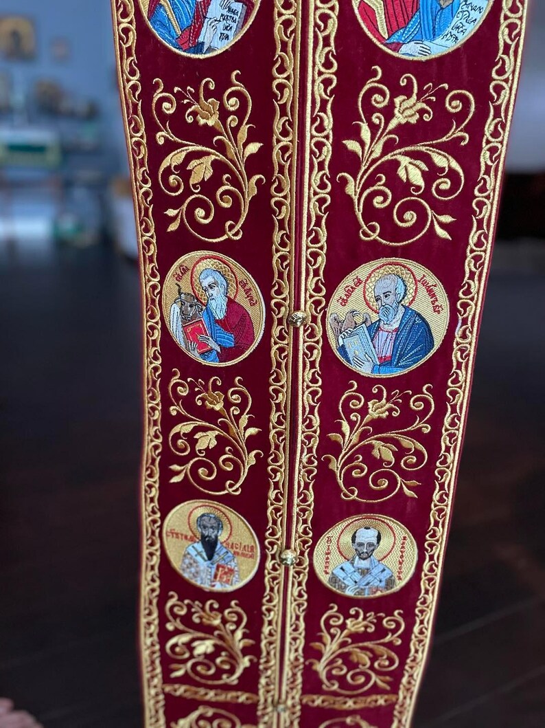 Pode incluir: Um tecido de veludo vermelho com bordados dourados. O tecido apresenta um padr&atilde;o repetitivo de desenhos florais e imagens circulares de figuras religiosas. O texto "&Epsilon;ὐ&lambda;&omicron;&gamma;&eta;&tau;ὸ&sigmaf; ὁ &Theta;&epsilon;ὸ&sigmaf; ἡ&mu;ῶ&nu;" est&aacute; bordado em ouro no tecido.