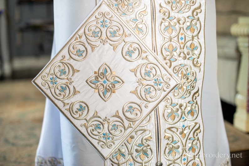 May include: White fabric with intricate gold and blue embroidery. The design features a repeating pattern of stylized flowers and leaves.