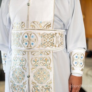 May include: White liturgical robe with gold and blue embroidery. The robe features a cross and the letters "RC" on the sash.