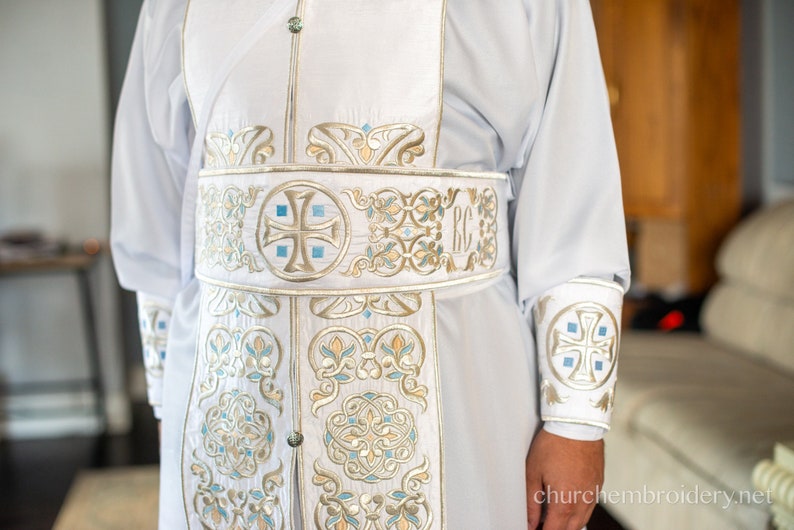 May include: White liturgical robe with gold and blue embroidery. The robe features a cross and the letters "RC" on the sash.
