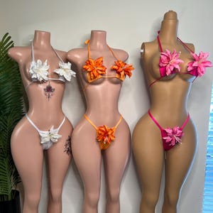 May include: Three floral bikini sets on mannequins. The sets feature white, orange, and pink tops and bottoms, each adorned with large, matching flower accents. The bikinis are displayed on mannequins with tan skin tones.
