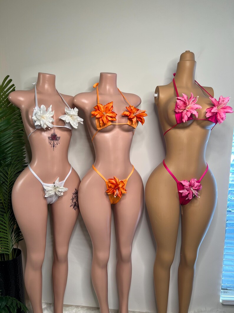 May include: Three floral bikini sets on mannequins. The sets feature white, orange, and pink tops and bottoms, each adorned with large, matching flower accents. The bikinis are displayed on mannequins with tan skin tones.