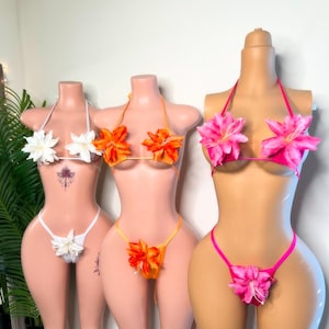 May include: Three floral bikini sets on mannequins. The sets feature flower accents on the tops and bottoms. One is white, one is orange, and the other is pink. The mannequins are displayed against a neutral background.