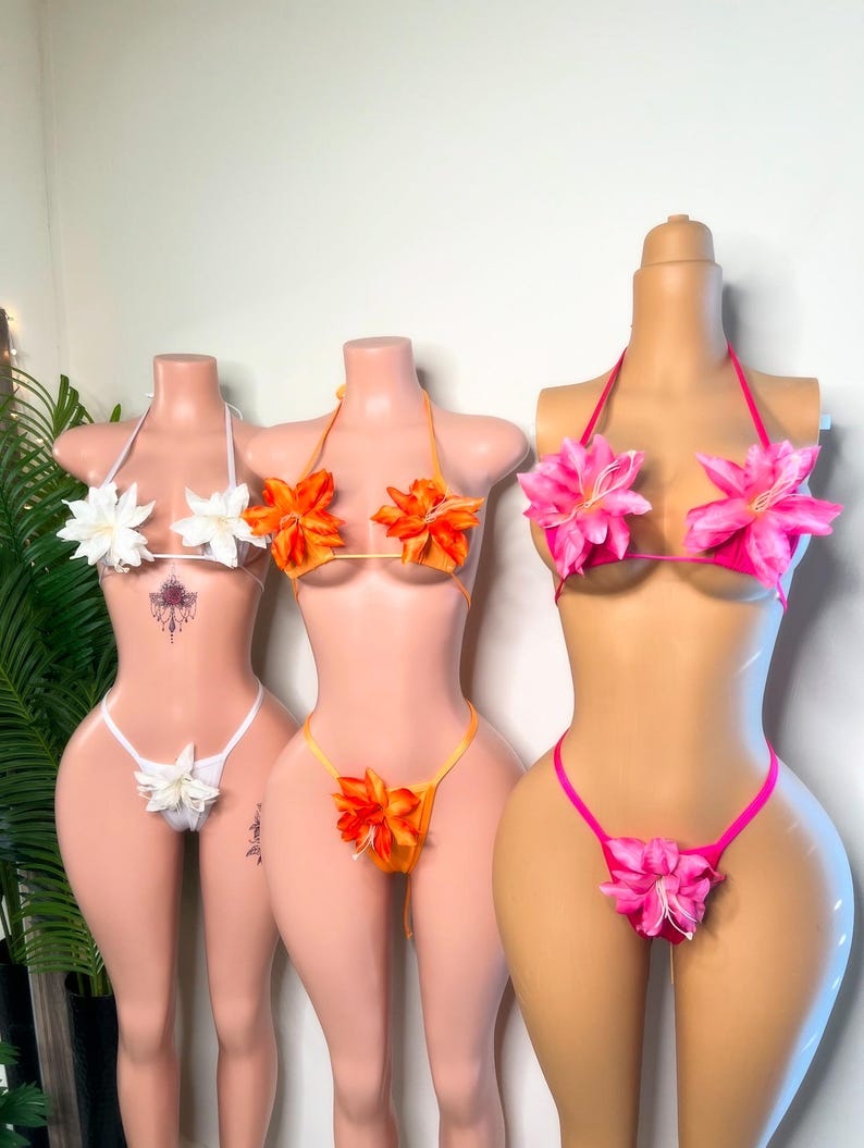 May include: Three floral bikini sets on mannequins. The sets feature flower accents on the tops and bottoms. One is white, one is orange, and the other is pink. The mannequins are displayed against a neutral background.