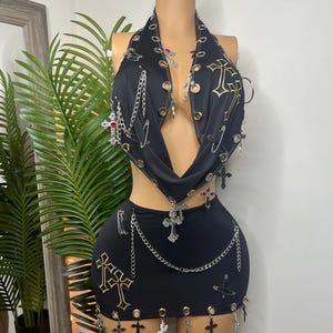 May include: Black two-piece outfit featuring a halter top and mini skirt. The top and skirt are adorned with gold crosses, silver chains, and cross charms. The outfit has grommet details and is displayed on a mannequin.