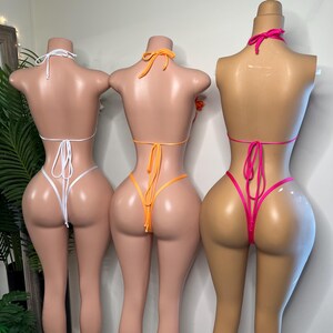 May include: Three mannequins display colorful bikinis. The first mannequin wears a white bikini, the second an orange bikini, and the third a hot pink bikini. Each bikini features a halter top and a thong bottom, tied with strings.