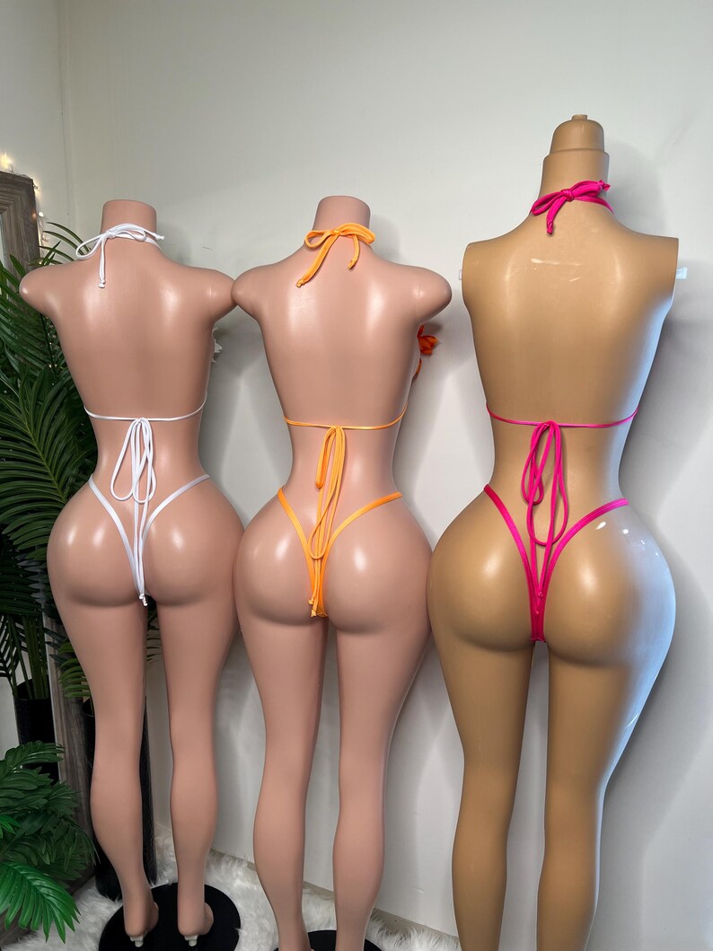 May include: Three mannequins display colorful bikinis. The first mannequin wears a white bikini, the second an orange bikini, and the third a hot pink bikini. Each bikini features a halter top and a thong bottom, tied with strings.