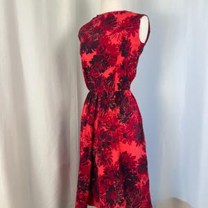 May include: A sleeveless red dress featuring a floral design in shades of red and dark purple. The dress has a fitted waist and a knee-length skirt. The dress is displayed on a mannequin against a white backdrop.