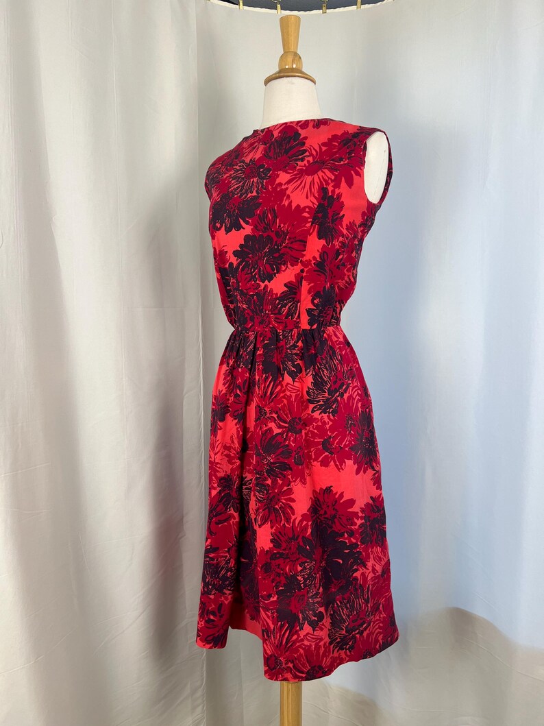May include: A sleeveless red dress featuring a floral design in shades of red and dark purple. The dress has a fitted waist and a knee-length skirt. The dress is displayed on a mannequin against a white backdrop.
