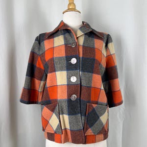 May include: A vintage plaid jacket in shades of orange, brown, gray, and beige. The jacket features a classic collar, three large buttons, and two front pockets. The sleeves are three-quarter length.