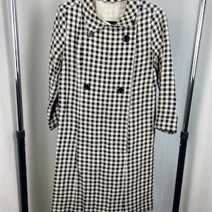 May include: A long-length double-breasted coat featuring a black and white gingham check pattern. The coat includes a rounded collar, long sleeves, and four black buttons arranged in two vertical rows on the front of the garment.
