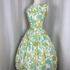 Puede incluir: Vestido vintage sin mangas con estampado floral. El vestido presenta un fondo blanco con un patrón repetido de flores y hojas amarillas, celeste y verdes. Tiene la cintura entallada y una falda amplia.