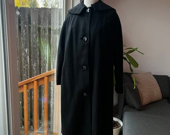 May include: A long black coat with a collar and button fastening. The coat is on a mannequin and is displayed in front of a window.