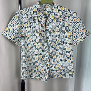May include: A short-sleeved button-down shirt with a white background and a repeating geometric pattern in green, yellow, orange, and red. The shirt has a collared neckline and is hanging on a green hanger.