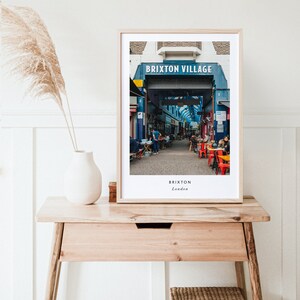 May include: A framed photograph of a street market in Brixton Village, London. The sign above the entrance reads "Brixton Village". The photo shows a bustling market with people walking through the stalls and sitting at tables.