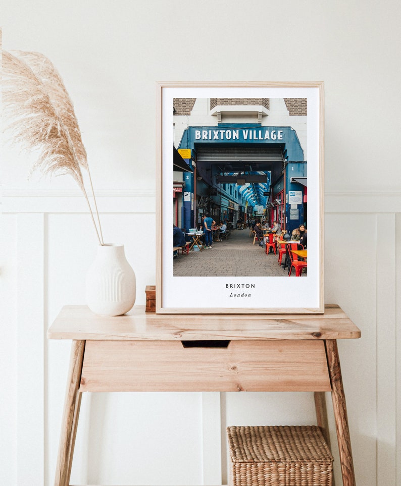 May include: A framed photograph of a street market in Brixton Village, London. The sign above the entrance reads "Brixton Village". The photo shows a bustling market with people walking through the stalls and sitting at tables.