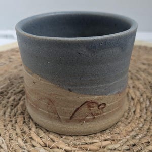 May include: A handmade ceramic mug with a two-tone design. The top half is a muted blue-grey, while the bottom half is a tan colour with a brown abstract design. The mug has a cylindrical shape and a smooth finish.