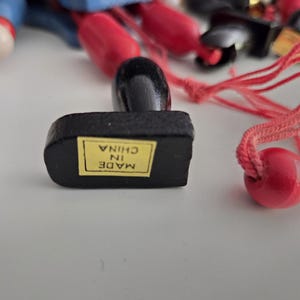 Peut inclure: Une base en bois noir avec un autocollant rectangulaire jaune portant l'inscription MADE IN CHINA. Un bouton en bois noir et une ficelle rouge avec une perle en bois rouge sont fix&eacute;s &agrave; la base. D'autres &eacute;l&eacute;ments en bois flous sont visibles.