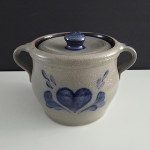 May include: A stoneware lidded jar with two handles. The jar is light gray with a dark blue heart and leaf design. The lid and knob are dark blue. The jar is likely used for food storage.