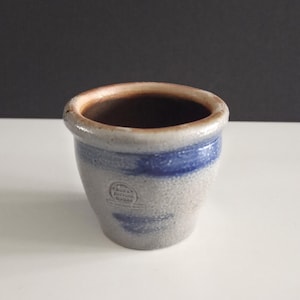 May include: A small, gray stoneware pot with a blue band around the middle. The rim is a darker brown color. The pot has a slightly textured surface and a stamped design on the front.