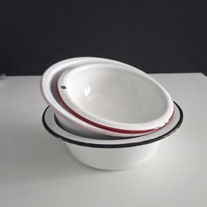 May include: Two white enamel bowls stacked on top of each other. The top bowl has a red rim, and the bottom bowl has a black rim. The bowls are on a white surface with a dark background.