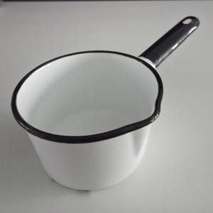 May include: A white enamel saucepan with a black handle and rim. The small saucepan has a pouring spout and is designed for heating liquids or small portions of food. The handle has a hole for hanging.