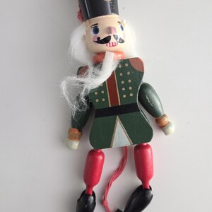 Peut inclure: Une figurine casse-noisette en bois avec un chapeau noir, des cheveux synth&eacute;tiques blancs et une veste verte peinte avec des d&eacute;tails rouges et blancs. Le personnage a des cuisses rouges, des jambes noires et des pieds noirs, avec une ficelle rouge.