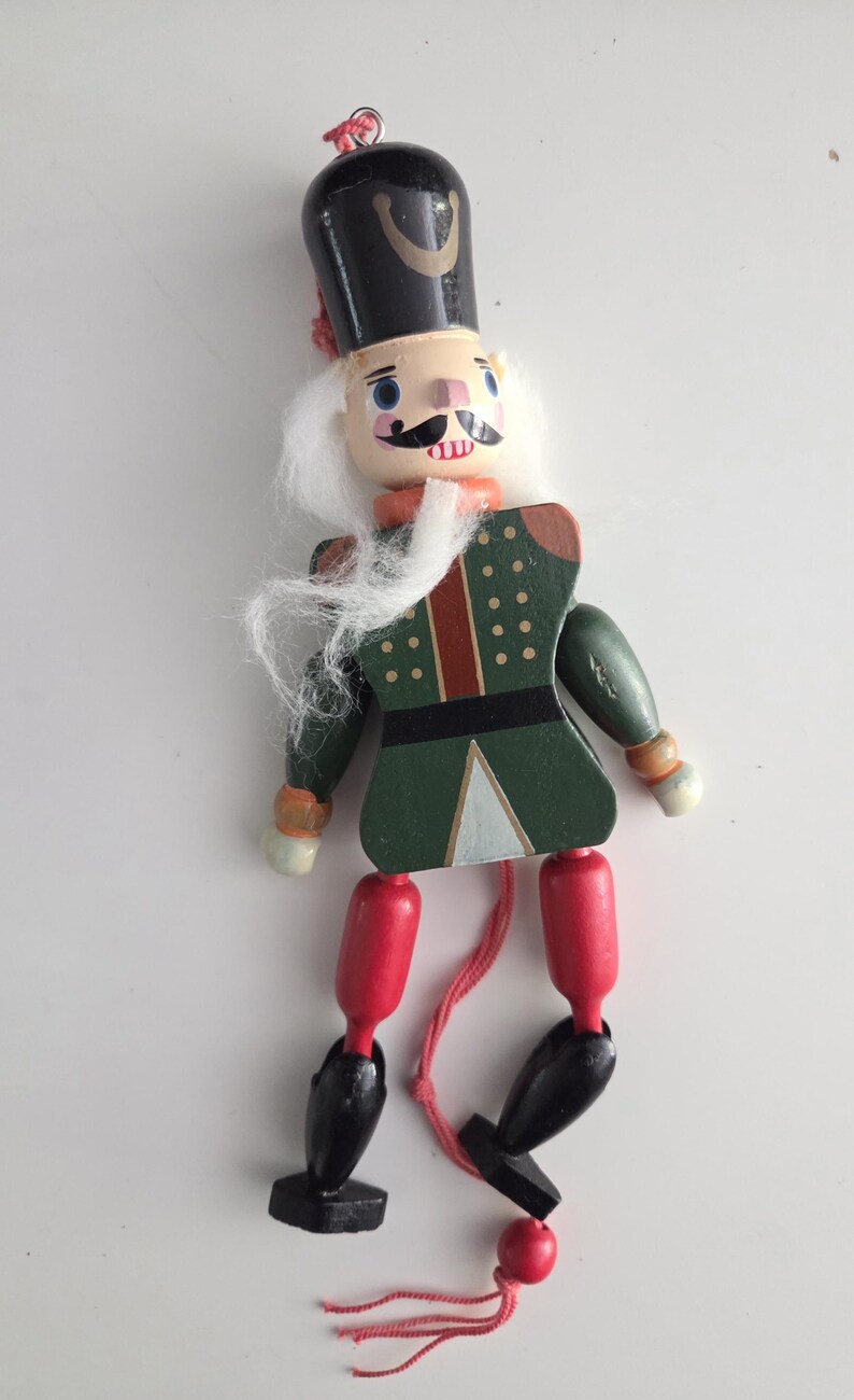 Peut inclure: Une figurine casse-noisette en bois avec un chapeau noir, des cheveux synth&eacute;tiques blancs et une veste verte peinte avec des d&eacute;tails rouges et blancs. Le personnage a des cuisses rouges, des jambes noires et des pieds noirs, avec une ficelle rouge.