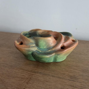 May include: A ceramic flower frog in shades of green, orange, and brown. The frog has a swirling, organic shape with multiple holes for arranging stems. The piece sits on a wooden surface.