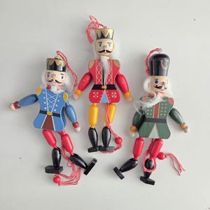 May include: Three wooden nutcracker ornaments with articulated limbs and red hanging strings. The left figure wears a blue jacket, the center figure a red jacket with a gold crown, and the right figure a green jacket with a black hat, all with white hair.