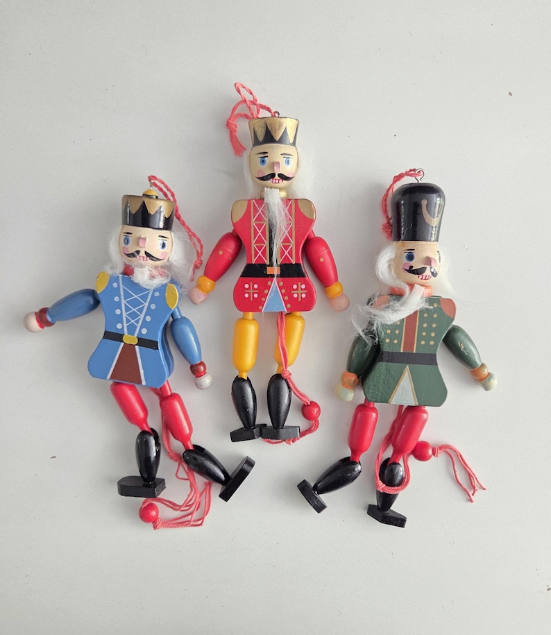 Peut inclure: Trois casse-noisettes en bois avec des membres articul&eacute;s et des cordons de suspension rouges. La figurine de gauche porte une veste bleue, celle du centre une veste rouge avec une couronne dor&eacute;e, et celle de droite une veste verte avec un chapeau noir.