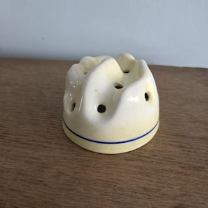 May include: A cream-colored ceramic kitchen item with a rounded, ridged top and multiple holes. A thin blue stripe encircles the base. The item is likely a kitchen tool for holding or separating items.