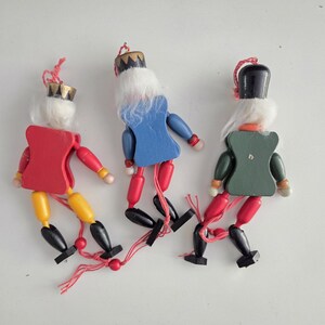 Peut inclure: Trois d&eacute;corations casse-noisettes en bois avec des cheveux blancs et des boucles en ficelle pour les suspendre. Les figurines ont des torses rouges, bleus et verts, des membres jaunes ou rouges et des bottes noires. Chaque pi&egrave;ce a une couronne ou un chapeau peint et des articulations reli&eacute;es par une ficelle rouge.