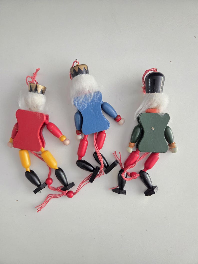 Peut inclure: Trois d&eacute;corations casse-noisettes en bois avec des cheveux blancs et des boucles en ficelle pour les suspendre. Les figurines ont des torses rouges, bleus et verts, des membres jaunes ou rouges et des bottes noires. Chaque pi&egrave;ce a une couronne ou un chapeau peint et des articulations reli&eacute;es par une ficelle rouge.