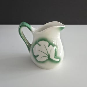 May include: A small, white ceramic creamer with a green handle and leaf design. The leaf is white with a green outline and shading. The creamer has a small spout for pouring.