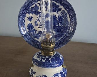 May include: A vintage oil lamp with a blue and white porcelain base and a clear glass chimney. The lamp features a decorative blue and white plate with a landscape design. The lamp is a classic piece of home decor.