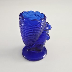 May include: A cobalt blue glass egg cup featuring a molded basket weave pattern on the exterior and a sculpted rabbit figure attached to the side. The vessel has a scalloped rim and stands on a circular base, crafted from translucent glass.