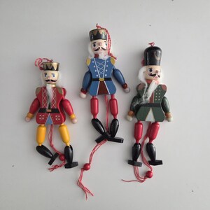 Peut inclure: Trois casse-noisettes en bois avec des membres articul&eacute;s et des ficelles de suspension. La figurine de gauche porte du rouge et du jaune, celle du centre du bleu, et celle de droite du vert et du rouge. Chaque personnage a une barbe blanche.
