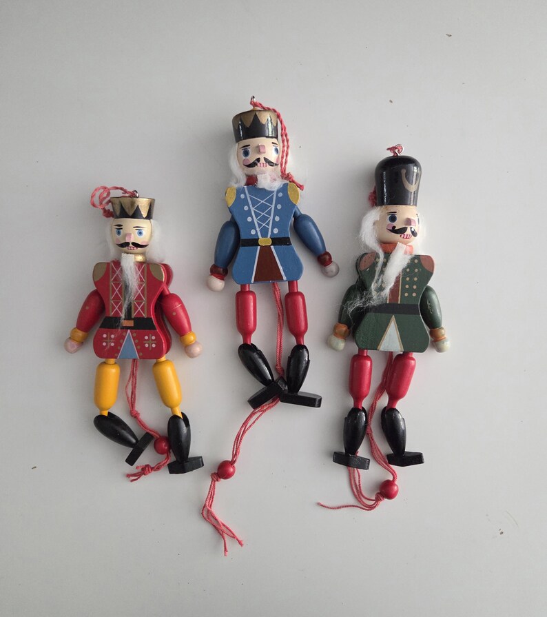 Peut inclure: Trois casse-noisettes en bois avec des membres articul&eacute;s et des ficelles de suspension. La figurine de gauche porte du rouge et du jaune, celle du centre du bleu, et celle de droite du vert et du rouge. Chaque personnage a une barbe blanche.