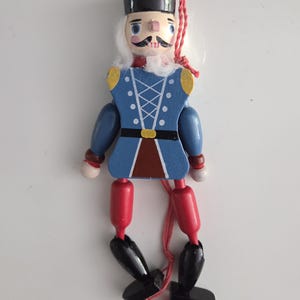 Peut inclure: Une d&eacute;coration casse-noisette en bois avec une couronne noire, des cheveux blancs et une veste bleue orn&eacute;e de d&eacute;tails de la&ccedil;age blancs. La figurine poss&egrave;de des cuisses rouges, des jambes noires et des pieds noirs, avec une ficelle rouge.