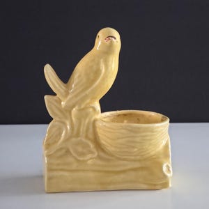 May include: A pale yellow ceramic bird figurine perched on a rectangular base with a small bowl. The bird has a detailed design with a red accent on its beak. The base has a leaf and wood-like texture.