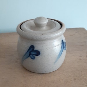 May include: A small, beige ceramic jar with a lid. The jar has a rounded shape and is decorated with blue floral designs. The lid has a small knob on top. The jar is sitting on a wooden surface.