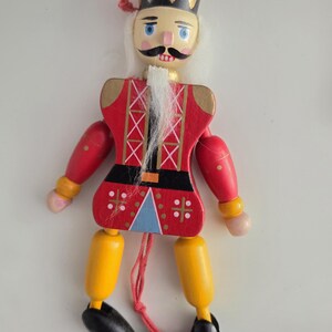 Peut inclure: Une marionnette casse-noisette en bois avec une couronne dor&eacute;e, une moustache noire et une barbe blanche. La figurine porte une veste rouge &agrave; croisillons blancs, une ceinture noire, des jambes jaunes et des chaussures noires, suspendue par un fil rouge.