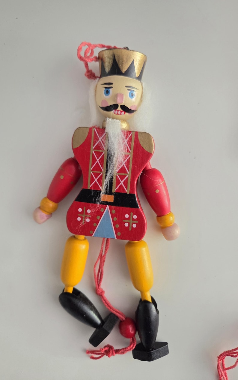 Peut inclure: Une marionnette casse-noisette en bois avec une couronne dor&eacute;e, une moustache noire et une barbe blanche. La figurine porte une veste rouge &agrave; croisillons blancs, une ceinture noire, des jambes jaunes et des chaussures noires, suspendue par un fil rouge.